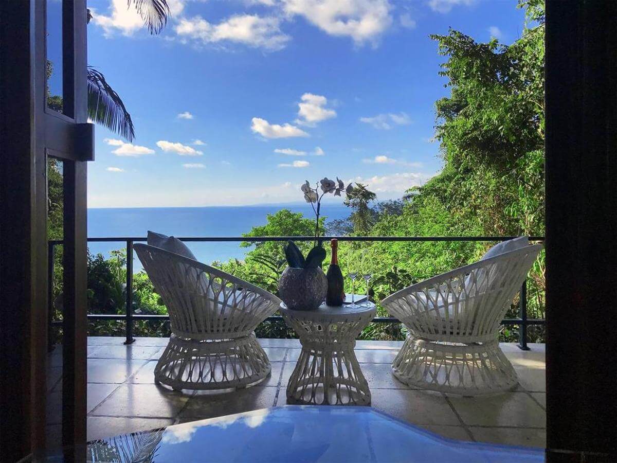 Private balcony with elegant white wicker chairs overlooking lush greenery and the Pacific Ocean. 