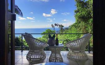 Private balcony with elegant white wicker chairs overlooking lush greenery and the Pacific Ocean. 