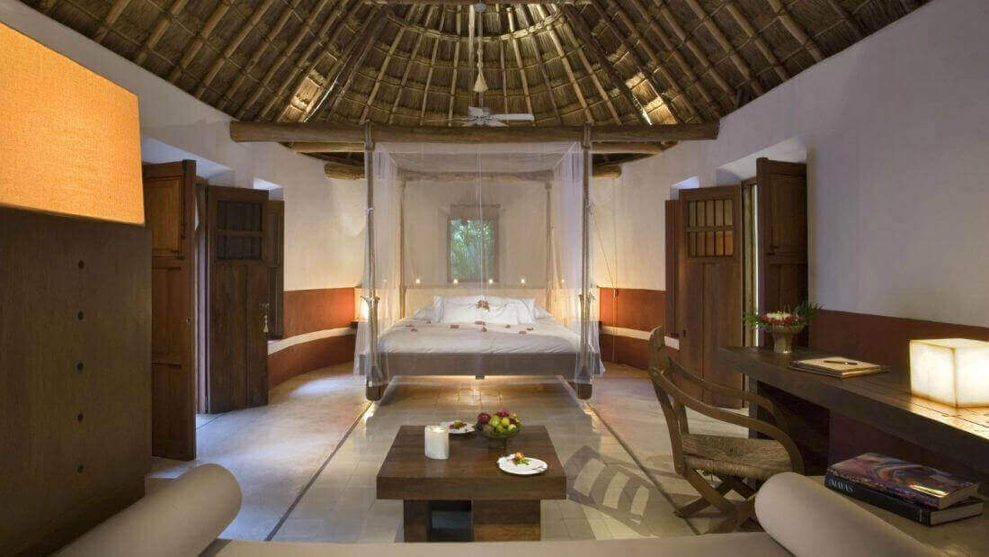 Spacious interior of a luxury Mayan-style villa with palapa roof, canopy bed, and natural wood furnishings in Mexico. 