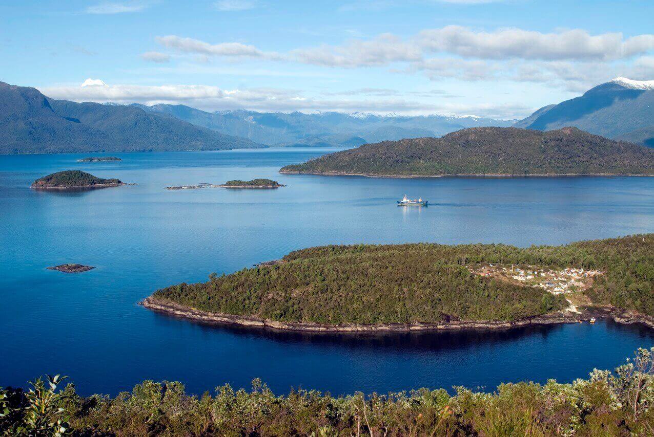 A cruise ship navigates the deep blue waters of Chile's fjords, weaving between lush green islands and distant Andean peaks. 