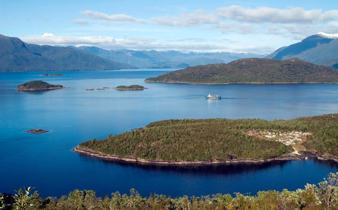 A cruise ship navigates the deep blue waters of Chile's fjords, weaving between lush green islands and distant Andean peaks. 