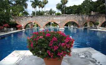 Charming Mexican hacienda with a serene pool, stone arches, and vibrant flowers in a tranquil garden setting. 