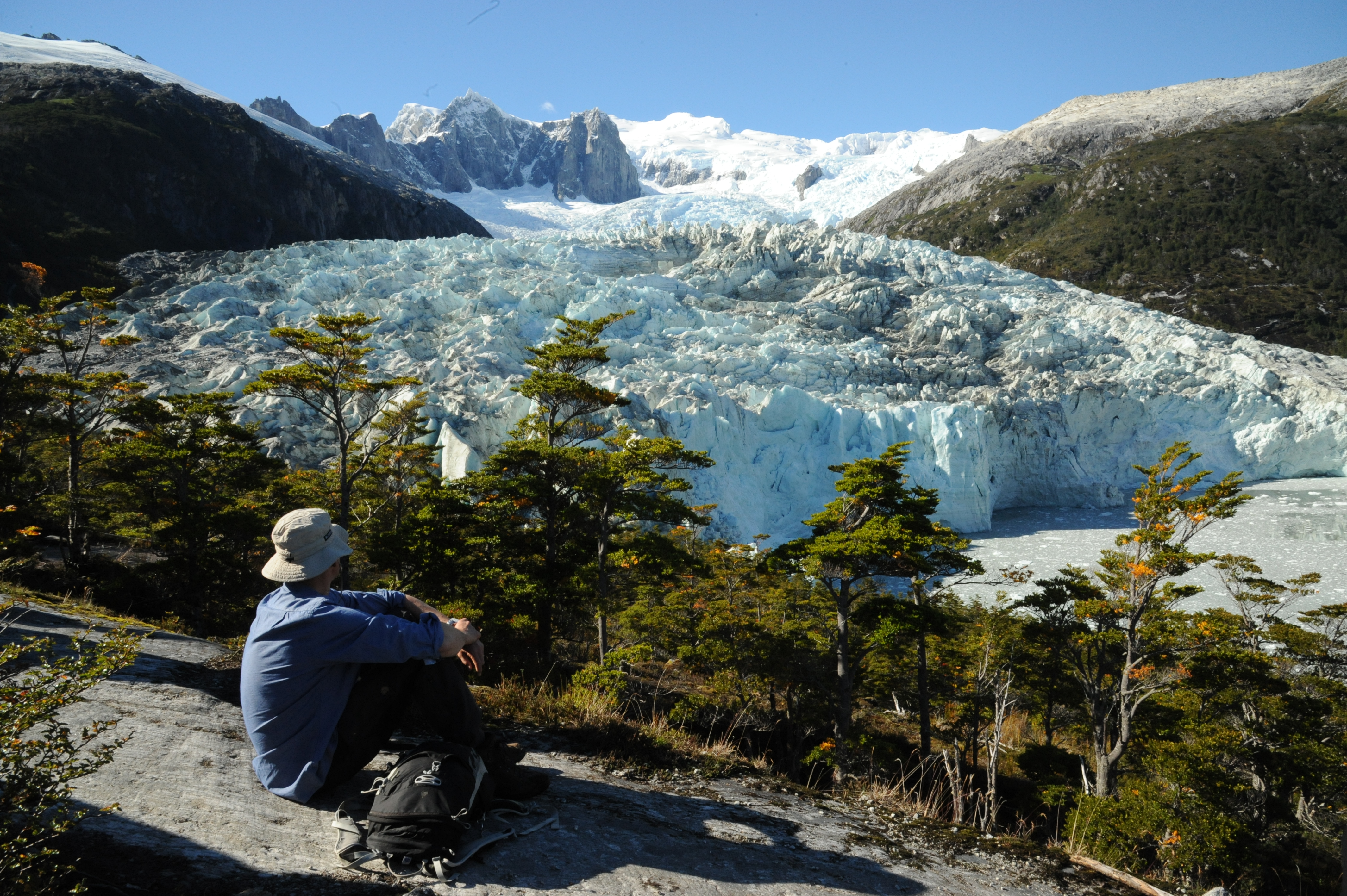 A lone hiker rests on a rock ledge, gazing at a massive glacier framed by snowy peaks in Chilean Patagonia. 