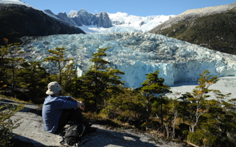 A lone hiker rests on a rock ledge, gazing at a massive glacier framed by snowy peaks in Chilean Patagonia. 