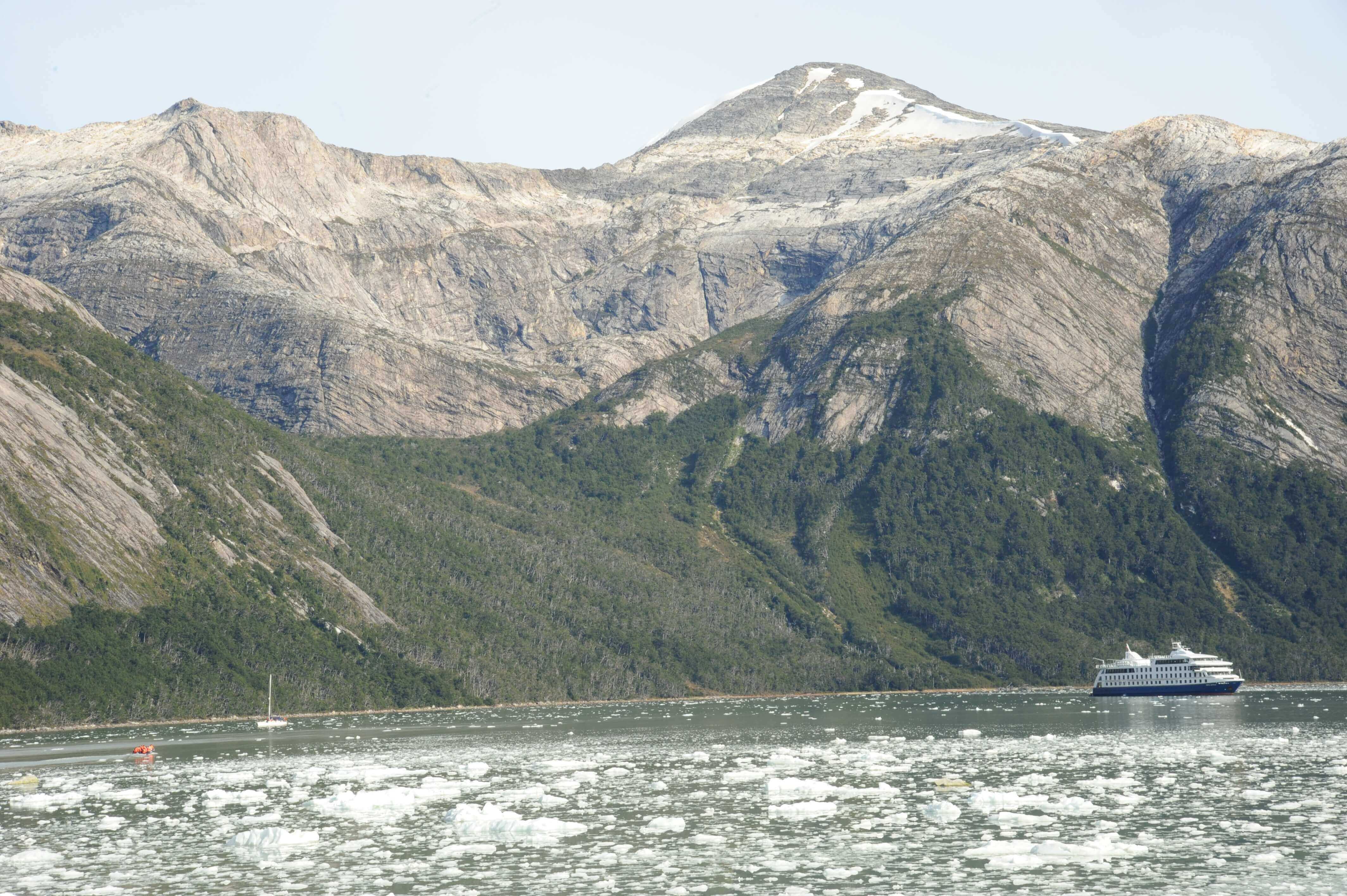 An expedition cruise ship navigates icy Patagonia waters beneath towering, rugged mountains in Chile's southern fjords. 