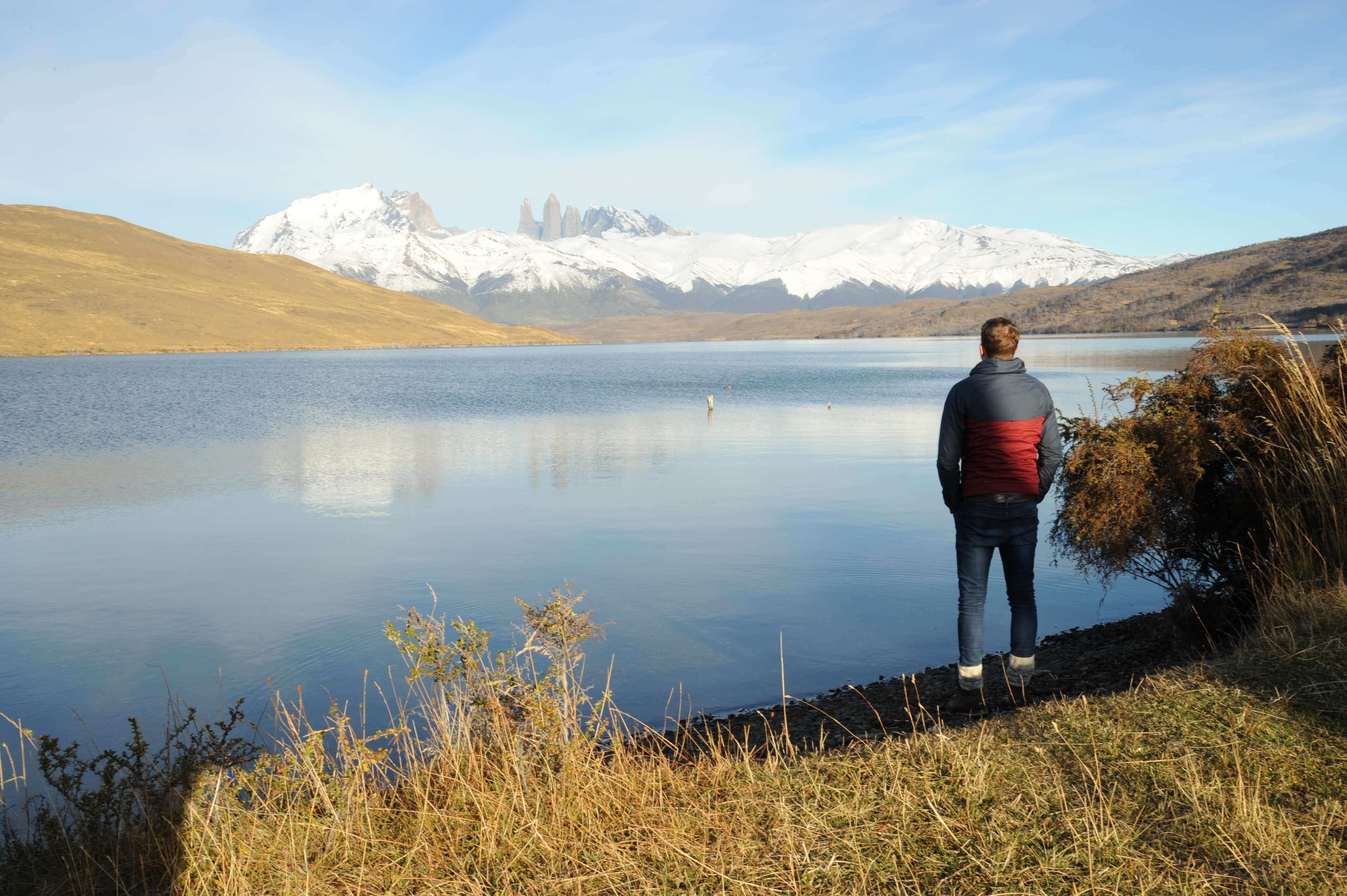 A lone visitor stands beside a still lake reflecting snow-capped peaks in Chile's Torres del Paine National Park. 