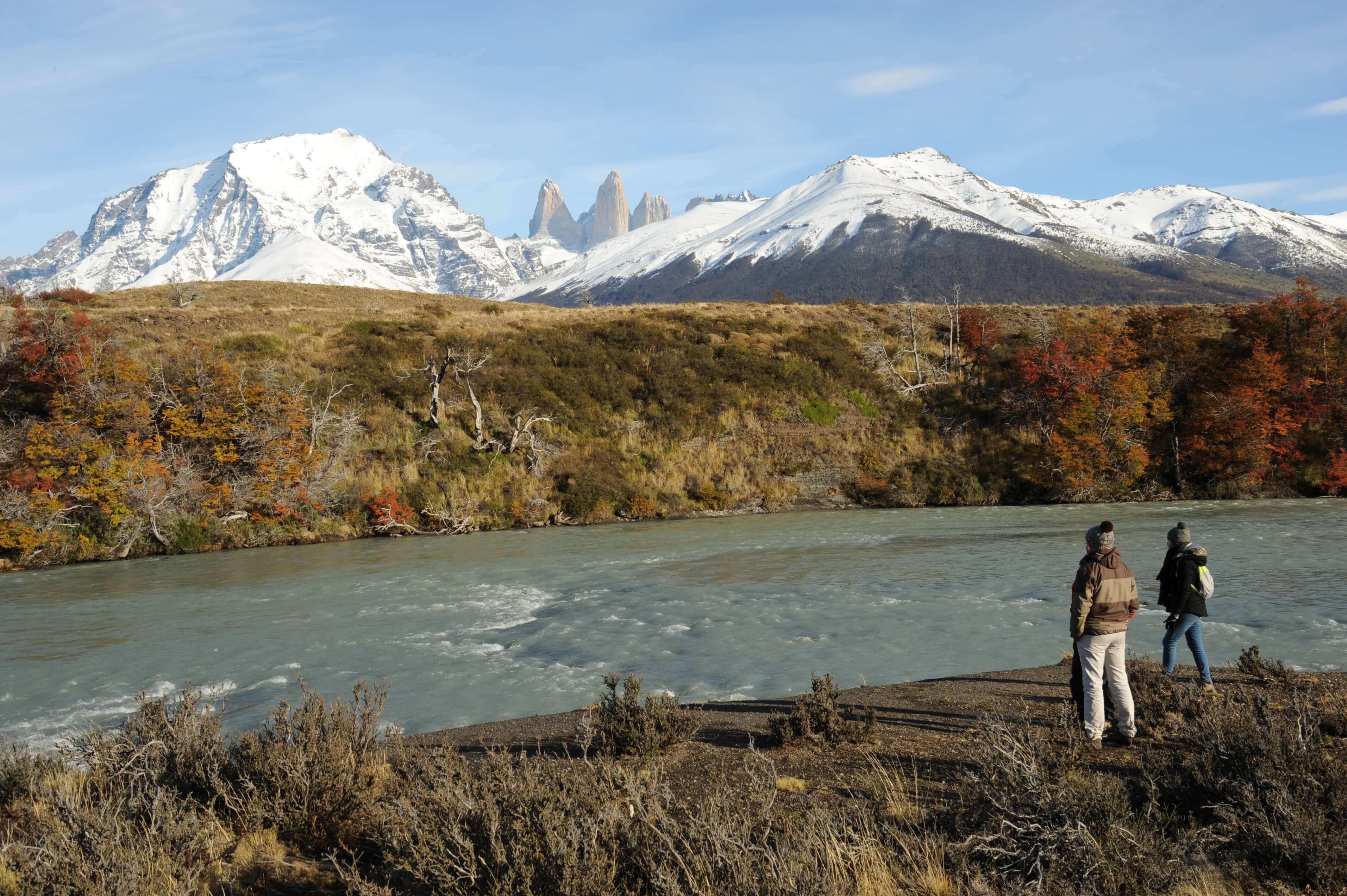 Two Hikers stand by a glacial river with Torres del Paine's snow-capped peaks towering in the background in Chilean Patagonia. 