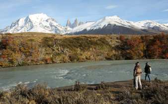 Two Hikers stand by a glacial river with Torres del Paine's snow-capped peaks towering in the background in Chilean Patagonia. 