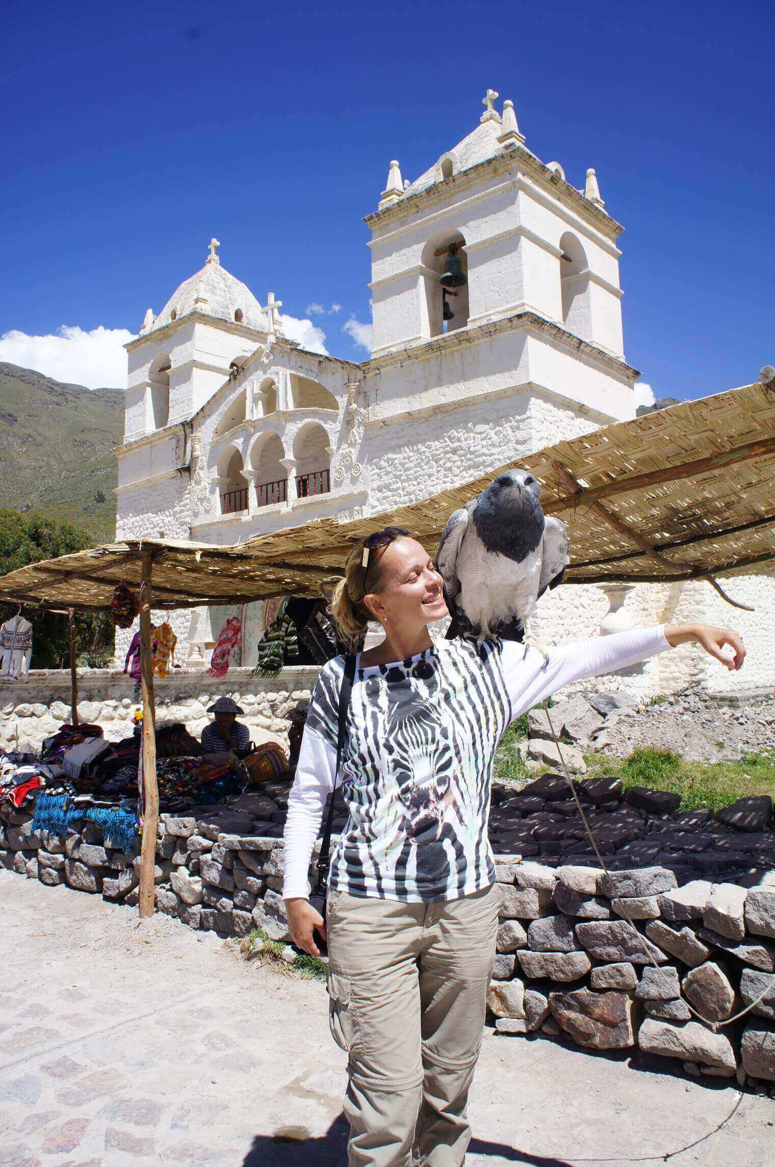 A traveler smiles with a parrot in Chivay's lively market, beneath the white towers of a historic Andean church. 