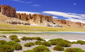 Dramatic rocks spires rise above green shrubs and a salt flat in Chile's high altitude Altiplano region. 