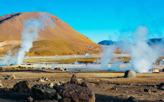 Steam rises from El Tatio geysers in Chile's Atacama Desert, surrounded by rocky terrain and golden morning light. 