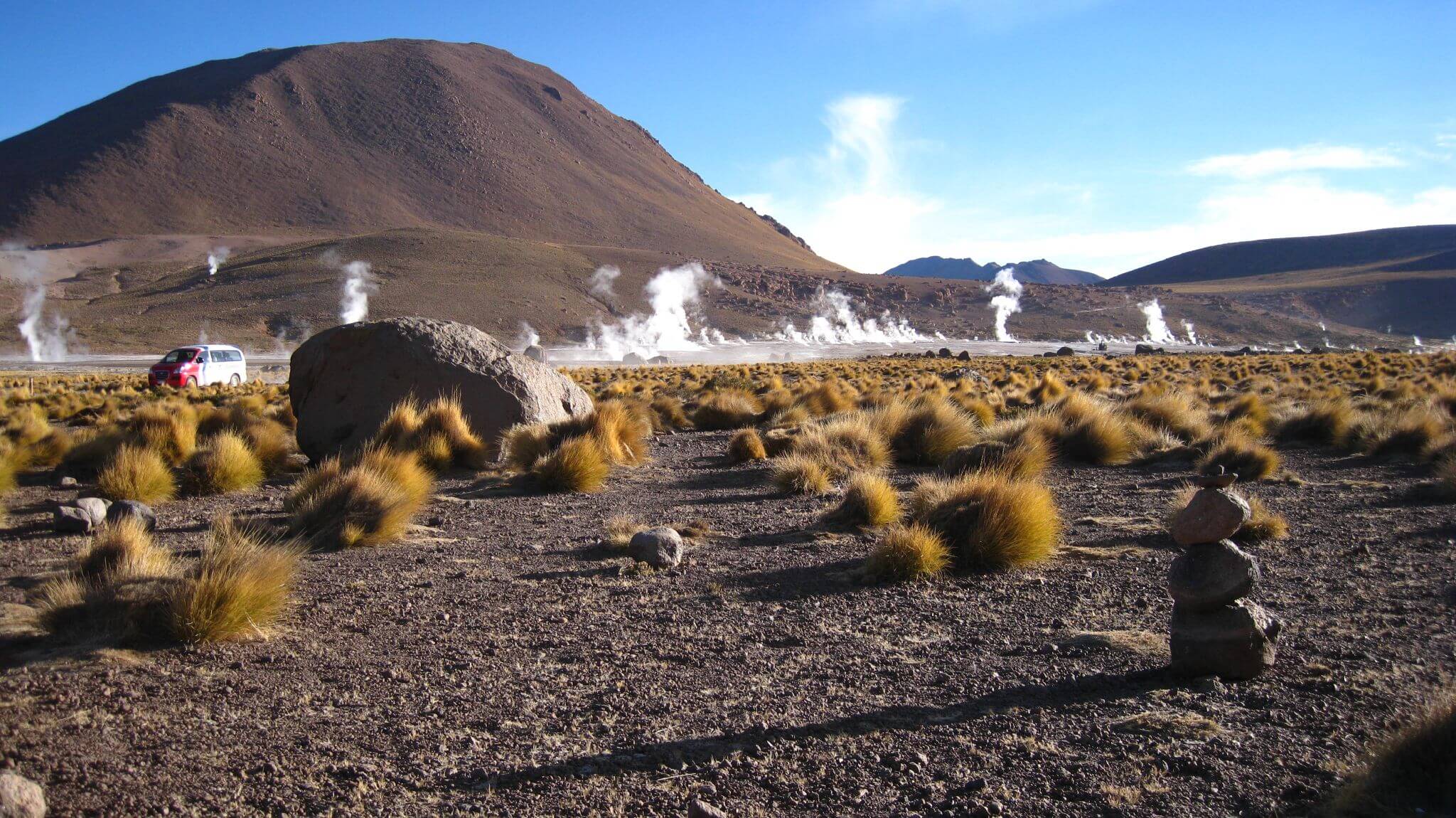 Steam rises from El Tatio geysers at sunrise, framed by desert vegetation and volcanic peaks. 