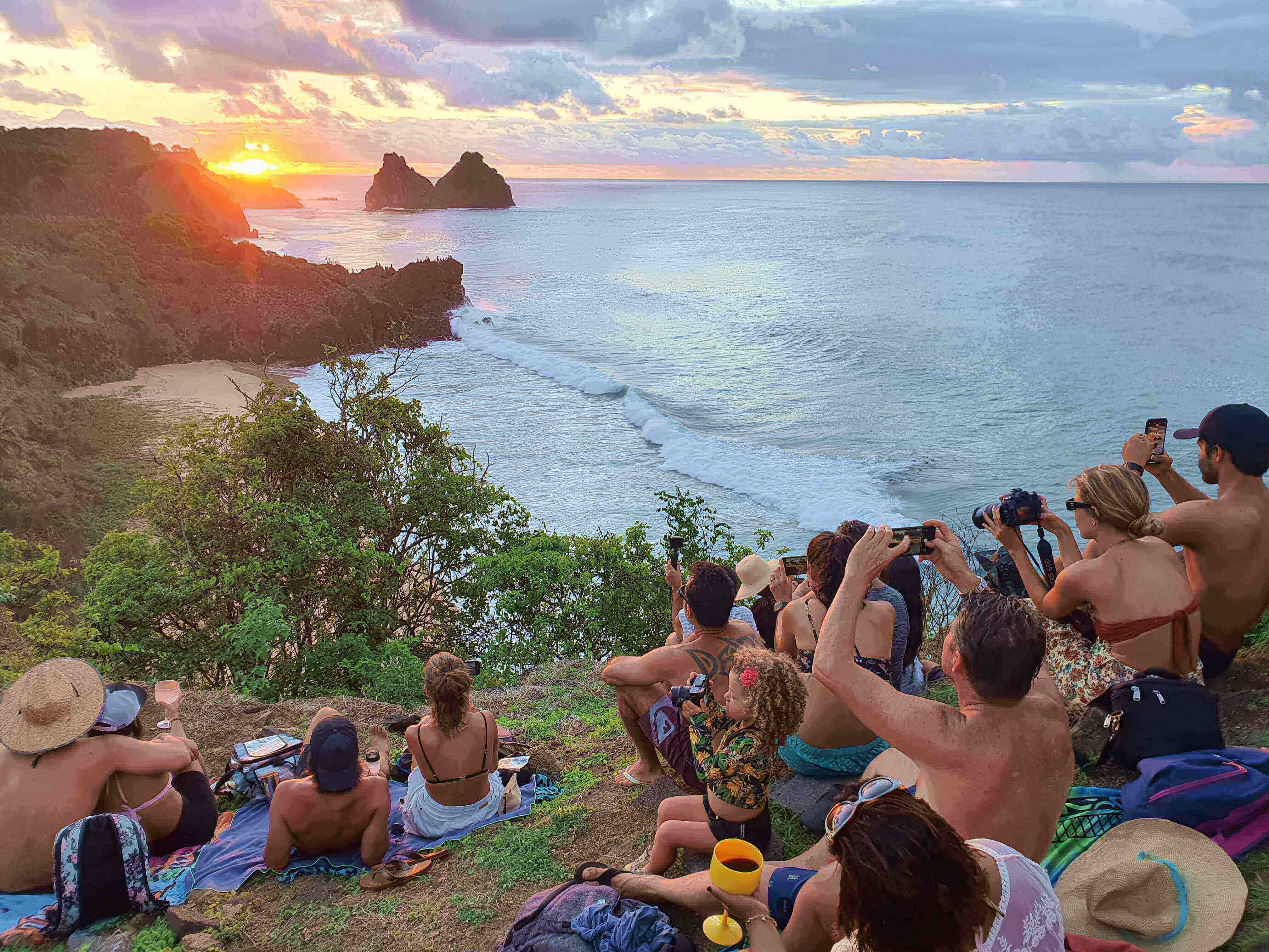 Tourists gathered at sunset viewpoint overlooking the ocean and Morro Dois Irmaos rocks in Fernando de Noronha, Brazil. 