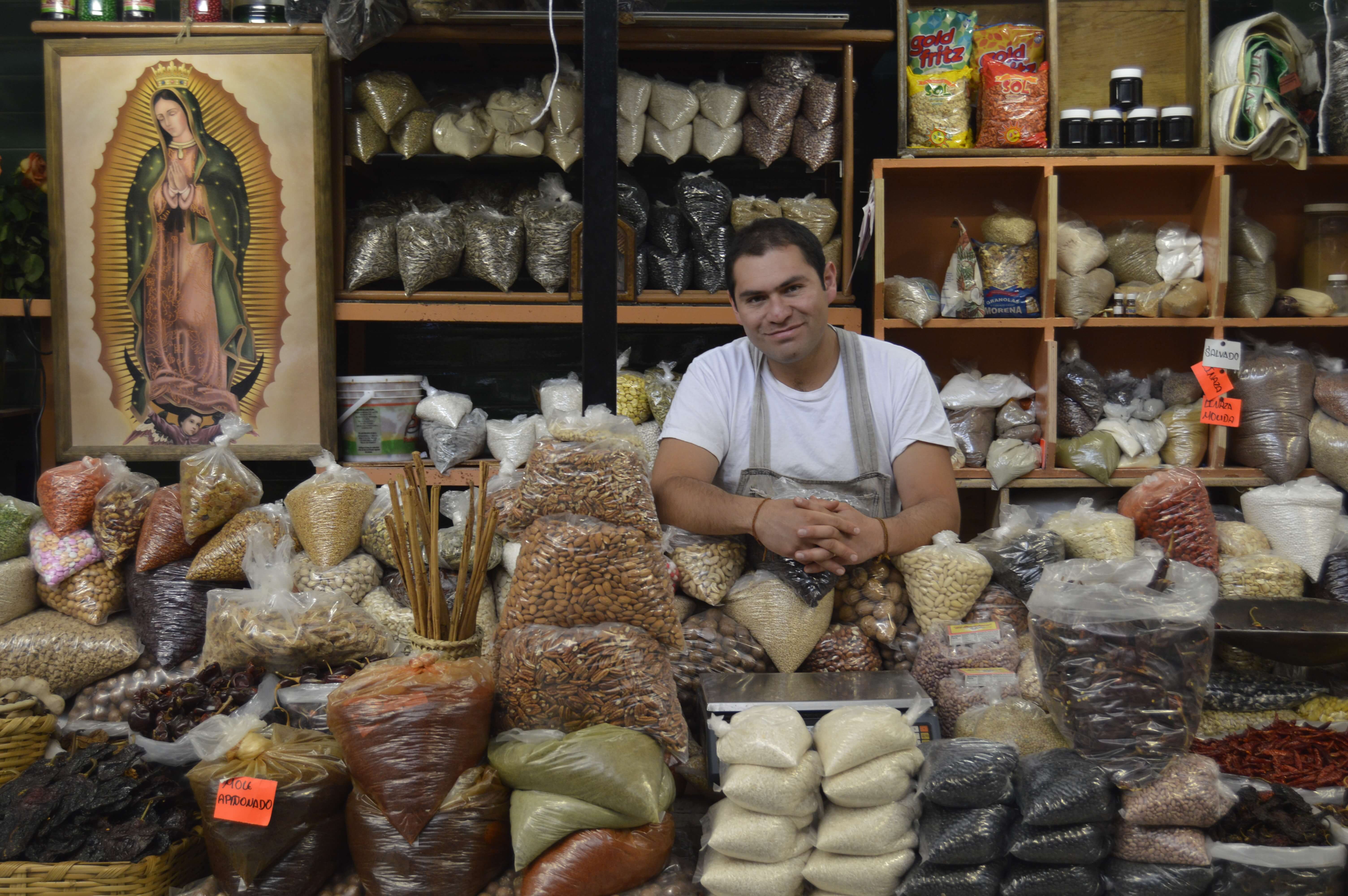 Friendly vendor in San Miguel de Allende sits among sacks of grains and spices, under the gaze of the Virgin of Guadalupe. 